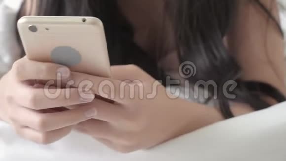 美丽的亚洲年轻女性看着摸着智能手机在卧室的床上享受社交网络视频的预览图