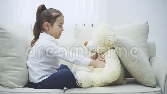 迷人的孩子在争吵后抱着她柔软的泰迪坐在沙发上视频的预览图