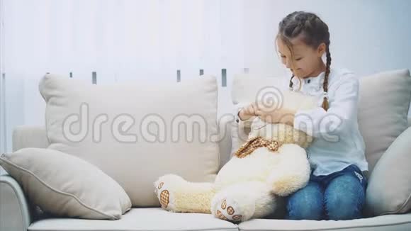 漂亮的女孩坐在沙发上玩着她的泰迪熊摸着它的鼻子和耳朵视频的预览图