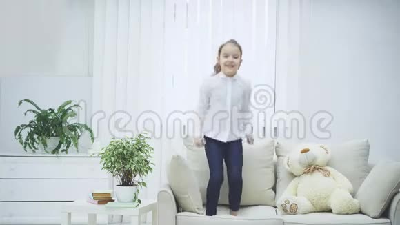 开朗的小女孩正欢快地在沙发上蹦蹦跳跳笑眯眯的她身边柔软的泰迪熊视频的预览图