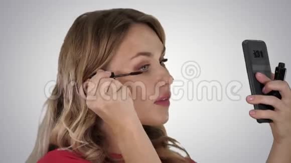 漂亮的金发年轻女人涂睫毛膏看着梯度背景上的智能手机镜子视频的预览图