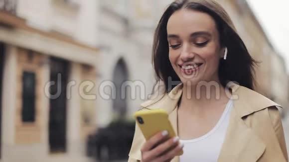 靠近年轻的微笑美丽的高加索女孩戴着耳机走在街上手里拿着电话视频的预览图