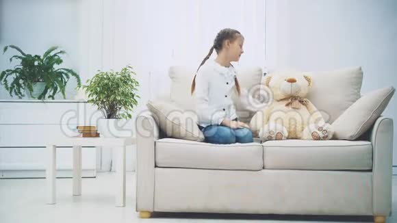 可爱迷人的孩子正坐在沙发上她的腿蜷缩着和旁边的人说话白色泰迪熊视频的预览图
