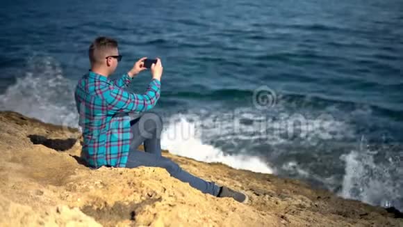 一个年轻人坐在海前的石头上拿着电话戴墨镜的人拍摄大自然的照片溅起的视频的预览图