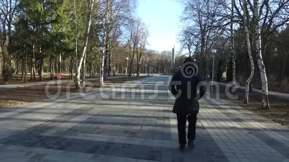 一个身穿黑色外套头罩的无名男子在秋天公园的小路上走来慢动作视频的预览图