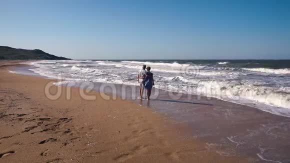一对苗条的夫妇一个穿着短裤的男人和一个穿着蓝色裙子的女人在沙滩上在强风中赤脚行走令人钦佩视频的预览图
