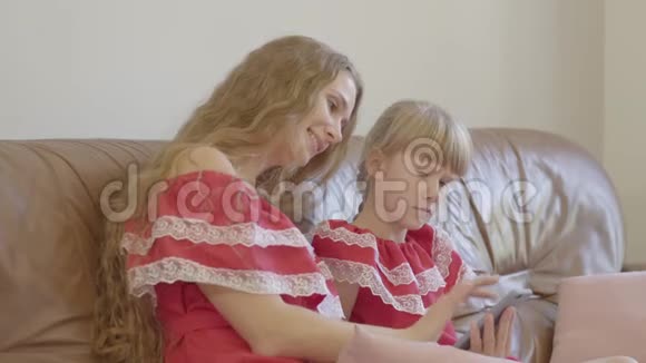 快乐的金发白种人母亲和小女儿穿着同样的衣服坐在沙发上在家里用手机概念视频的预览图