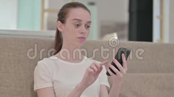 红发青年女性在沙发上使用智能手机视频的预览图