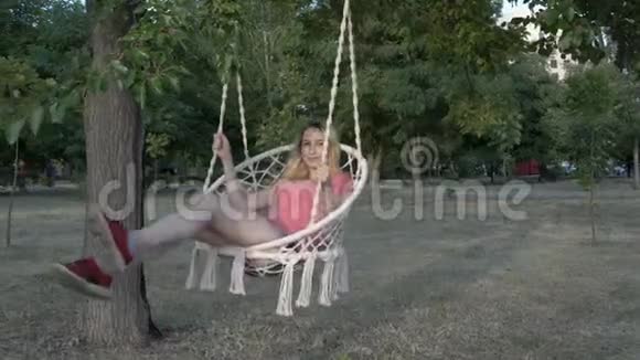 日落时分一个穿着粉色t恤的少女在城市公园里用绳子编织的秋千上荡秋千微笑着视频的预览图