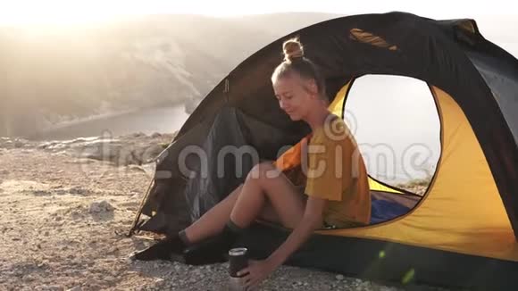 金发女孩露营者坐在靠近帐篷入口的山和湖的背景喝咖啡或茶视频的预览图