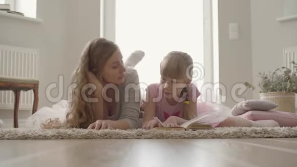 快乐的金发白种人母亲和小女儿躺在家里蓬松的地毯上女孩在看书视频的预览图