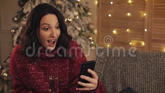 令人惊讶的年轻美女拿着一杯香槟坐在圣诞树旁的智能手机的画像视频的预览图