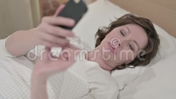 漂亮的老妇人在床上讲智能手机视频的预览图
