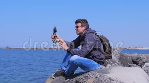 旅行博主man在智能手机上拍摄视频坐在海边的石头上视频的预览图