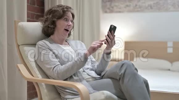 在沙发上庆祝智能手机成功的老妇人视频的预览图