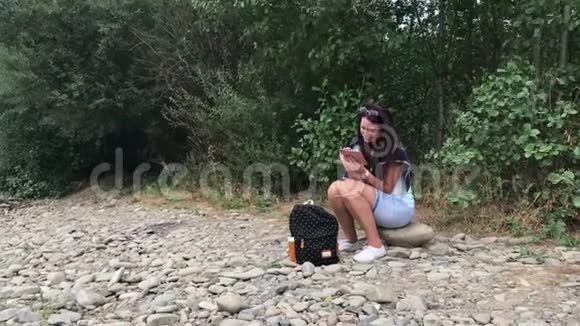 一个女孩坐在山河的岸边看着一部智能手机附近的旅游用品河的对岸视频的预览图