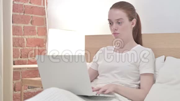 红发青年妇女在笔记本电脑上工作在床上头痛视频的预览图