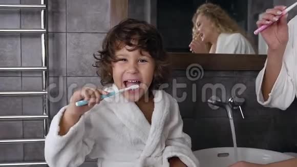 穿着白色外套的卷发开朗的小男孩正在用牙膏和牙刷一起刷牙视频的预览图