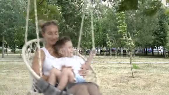 可爱的女孩和一个孩子在秋千上摆动挂在公园的一棵树上一个小男孩坐在妈妈腿上他们视频的预览图
