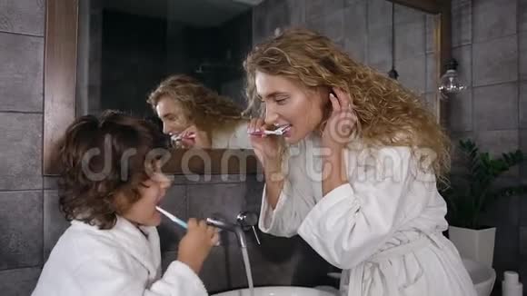 漂亮的年轻女人留着长长的卷发穿着白色浴袍和她可爱的儿子在早上的前面刷牙视频的预览图