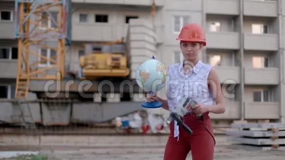 戴着钻和地球仪的建筑头盔的年轻女子在建筑中的房子和起重机的背景下跳舞视频的预览图
