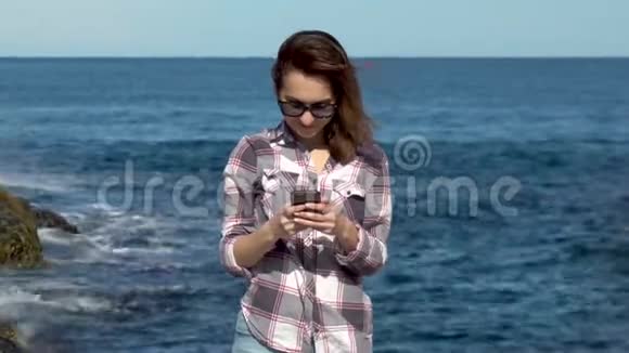 一个年轻的女人站在大海的背景上带着一个电话女人在度假时在智能手机上发短信阳光明媚视频的预览图