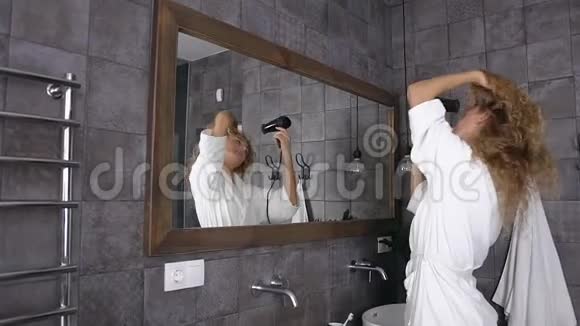 漂亮的金发女人穿着白色浴袍在家里的浴室里长发在镜子前晾晒头发视频的预览图