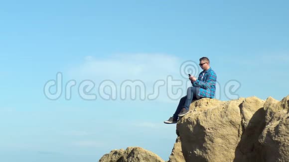 一个年轻人拿着电话坐在岩石上一个穿着蓝色衬衫的人把腿挂在悬崖上通信视频的预览图