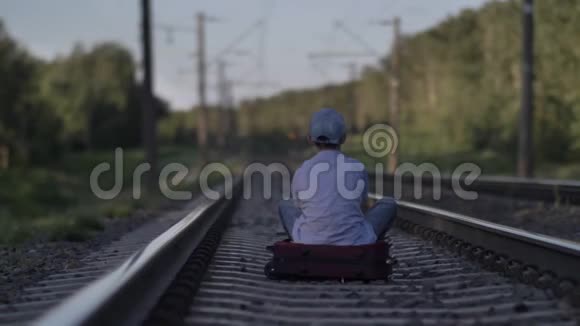 戴着帽子的悲伤男孩坐在铁路上的手提箱上看着室外的景色视频的预览图