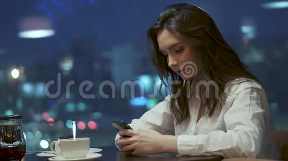年轻女孩正在智能手机上发短信坐在夜晚灯光背景下的咖啡馆里视频的预览图