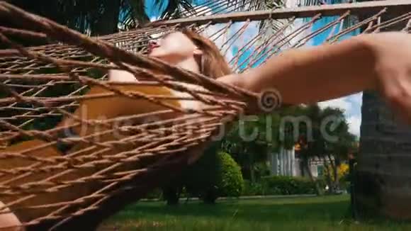 一个年轻快乐的女人躺在吊床上闭着眼睛看着热带植物休息一下视频的预览图