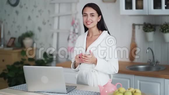 穿着白色浴袍的年轻女子在笔记本电脑上工作早上喝茶视频的预览图