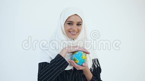 一位年轻快乐的穆斯林女性戴着头巾站在白色背景上用小地球仪在镜头上摆姿势视频的预览图
