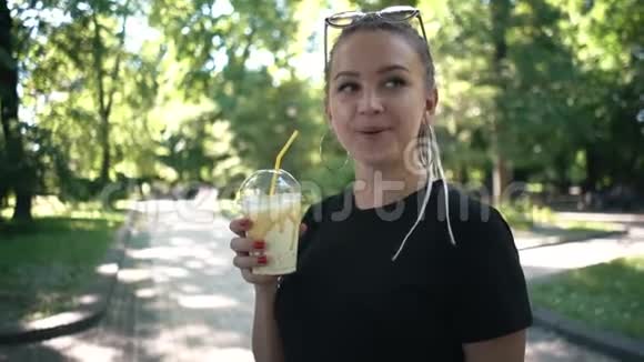 在阳光明媚的公园里年轻美丽的编辫子女人喝着焦糖奶昔视频的预览图
