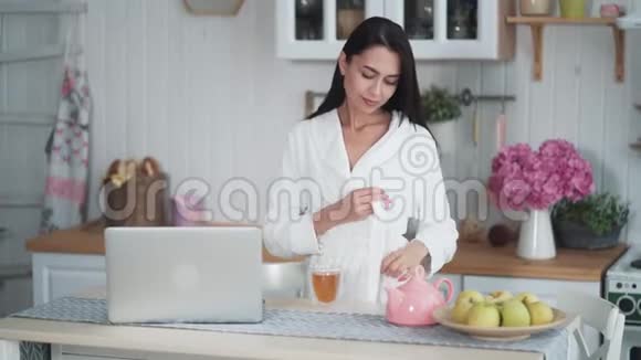 穿着白色浴袍的年轻女子在笔记本电脑上工作早上喝茶视频的预览图