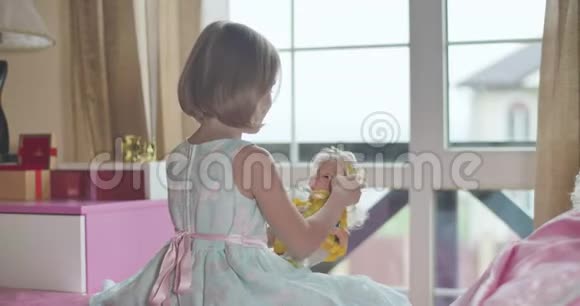 白人女孩穿着漂亮的蓝色连衣裙粉红色的蝴蝶坐在床上玩娃娃孩子抱着玩具视频的预览图