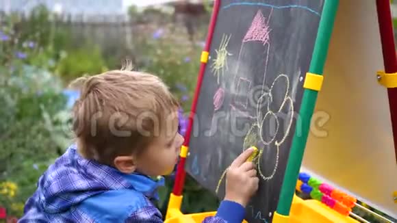 年轻的艺术家在黑板上画画一个男孩在开阔的空气中在鲜花和绿色植物中画孩子们的画视频的预览图