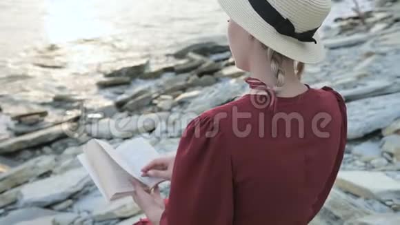 后景特写一位迷人的白种人女孩穿着红色鲜艳的连衣裙和一顶草帽坐在海边的一块大石头上视频的预览图