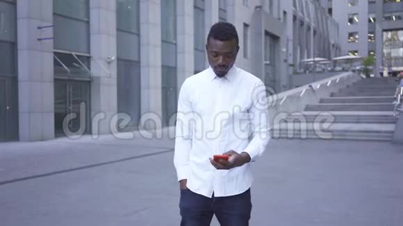 英俊的非洲裔美国人穿着白色衬衫站在城市街道上用红色手机说话男性商人或视频的预览图