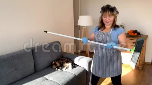 肥胖的中年妇女穿着围裙的家庭主妇用拖把用刷子在清洁房子的棍子上做体育锻炼视频的预览图