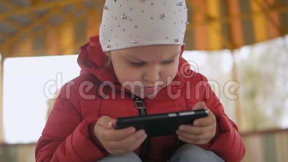 一个穿着红色夹克的小女孩专注地看着智能手机屏幕上的卡通视频的预览图