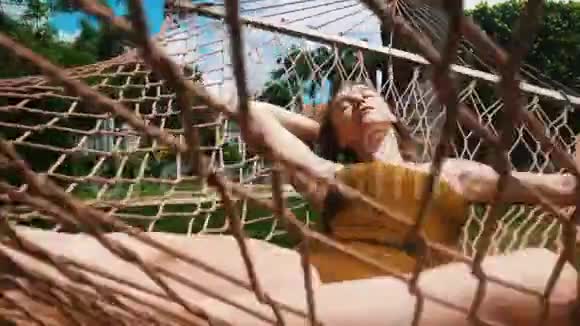 一位年轻漂亮的女士穿着一件芥末色泳衣躺在吊床上围着热带植物休息视频的预览图