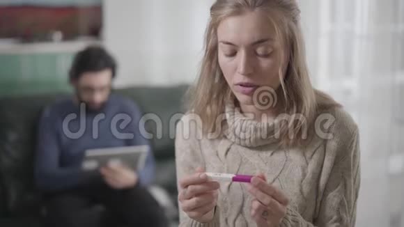 体贴的金发白种人女孩棕色的眼睛拿着怀孕测试回头看着男人用平板电脑看着视频的预览图