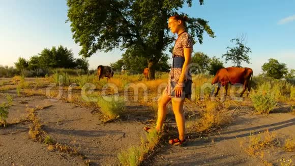 穿着短裙的牛仔女郎在夕阳下的牧场上远离镜头靠近长满沥青的炎热的乡村视频的预览图