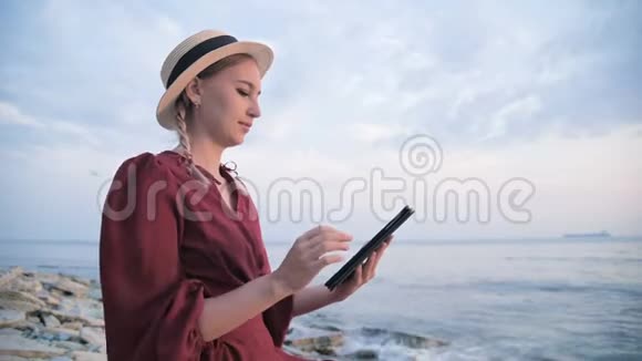 一个漂亮的女孩穿着一件夏天的红色连衣裙和一顶草帽晚上坐在海边的石头上视频的预览图