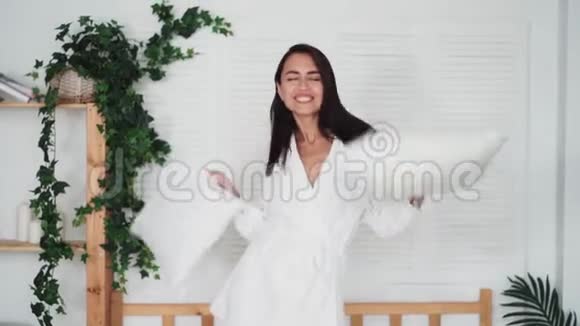 快乐的女人穿着浴袍带着枕头在床上跳跃动作缓慢有趣的视频视频的预览图