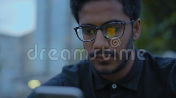 特写镜头戴眼镜的印度男人看着智能手机坐在公园的小巷里视频的预览图