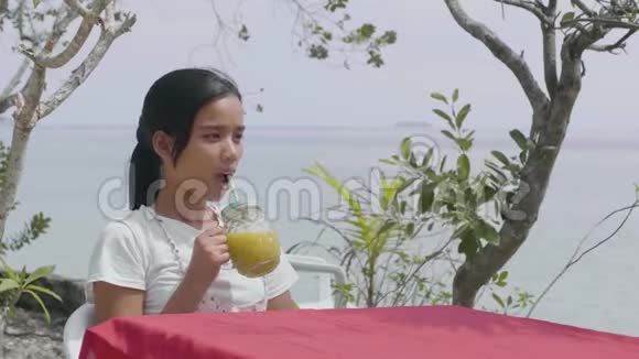 在热带海滨附近的餐桌上喝菠萝汁的漂亮年轻的亚洲女人视频的预览图