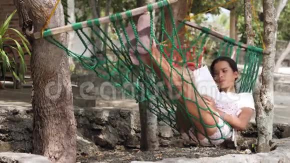 一个年轻的女人在户外树下的一个旋转吊床上看书视频的预览图
