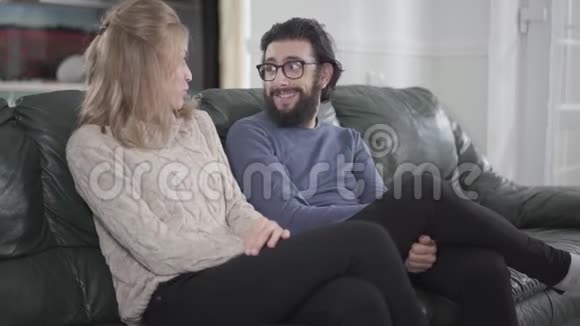 年轻的高加索夫妇坐在舒适的沙发上聊天戴眼镜的长胡子男人拥抱他的女朋友或视频的预览图